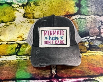 Może przedstawiać: Zniszczona czapka typu trucker w kolorze czarnym z białą siatką z tyłu. Na przedniej naszywce napis "MERMAID hair DON'T CARE" w różowym i niebieskim kolorze. Czapka ma zakrzywiony daszek i wyblakły wygląd.