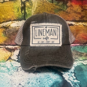 May include: A charcoal gray distressed trucker hat with a mesh back and a rectangular fabric patch on the front. The patch features the text LINEMAN WIFE with a small heart icon and the words ALL DAY EVERY DAY printed in black ink.
