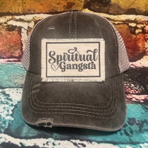 May include: A black and gray distressed trucker hat with a patch that says "Spiritual Gangsta" in black script with a heart symbol.