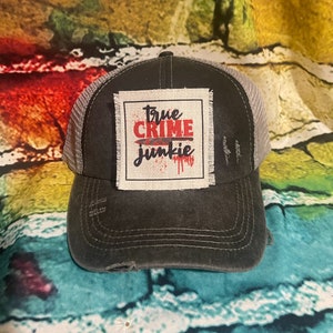 May include: A black and gray distressed trucker hat with a white patch that says "true crime junkie" in red letters with a red knife graphic and blood dripping.