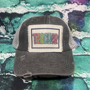 May include: A black and gray trucker hat with a distressed look. The hat has a patch on the front that says "TEACHER" in rainbow colors.