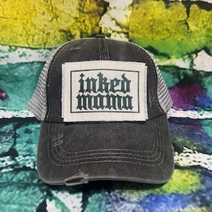 May include: A black and gray trucker hat with a white patch that says "inked mama" in green letters.