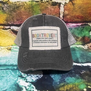 May include: A black and gray distressed trucker hat with a white patch that reads "BOOKTROVERT [book-tro-vert-noun] A person who prefers the company of fictional characters to real people."