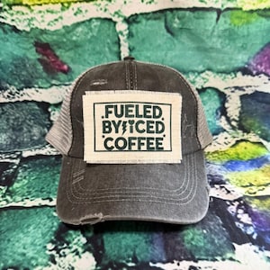 May include: A gray trucker hat with a mesh back and a distressed brim. The hat features a rectangular patch with the text "FUELED BY ICED COFFEE" in green and white. The hat is designed for casual wear.