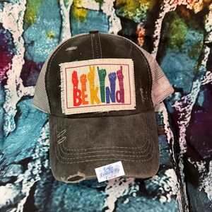 May include: A black distressed trucker hat with a beige patch featuring the word "BE KIND" in rainbow letters and colorful hand signs.