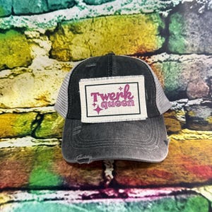 May include: A distressed black and gray trucker hat with a white mesh back. The hat features a rectangular patch with the words "Twerk queen" in pink, with a pink star graphic.