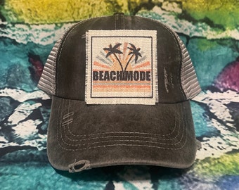 Może przedstawiać: Czarna, zniszczona czapka trucker z naszywką przedstawiającą dwie palmy i napis "Beach Mode" w kolorowym wzorze zachodu słońca.