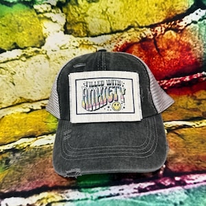 May include: A distressed black trucker hat with a white mesh back. The front patch reads "FILLED WITH: ANXIETY" in colorful text, with a smiling face. The hat has a pre-curved brim and a worn appearance.