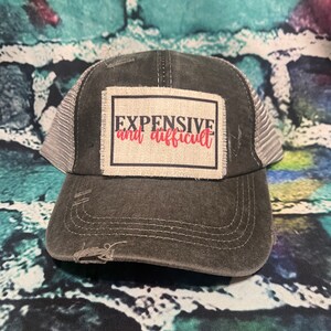 May include: A black and gray distressed trucker hat with a white patch that says "EXPENSIVE and difficult" in black and red lettering.