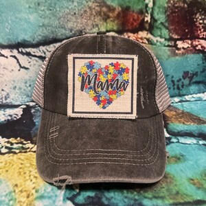 May include: A black distressed trucker hat with a gray mesh back. The hat features a patch with a heart made of puzzle pieces and the word "Mama" in a colorful font.