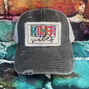 May include: A black and gray distressed trucker hat with a white patch that says "River Vibes" in rainbow letters.