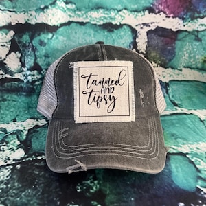 May include: A black and gray distressed trucker hat with a white patch that says "Tanned and Tipsy" in black script.
