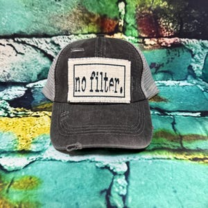 May include: A distressed charcoal gray trucker hat with a white mesh back. The front features a rectangular patch with the words "no filter" in black, outlined in a white border. The hat has a pre-curved brim.