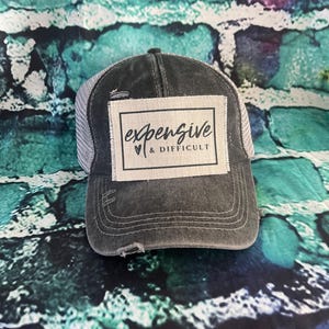 May include: A black and gray distressed trucker hat with a white patch that says "expensive & difficult" in black script.