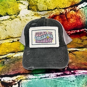 May include: A black trucker hat with a distressed brim and a white mesh back. The hat features a rectangular patch with the text "Fueled by Caffeine & Anxiety" in colorful, retro-style lettering. The hat is set against a colorful, textured background.