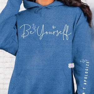 May include: A blue hoodie with the words "Be Yourself" in white script across the chest. The sleeve features the text "& DON'T APOLOGIZE" in white, along with a floral design. The hoodie has a front pocket and a drawstring hood.