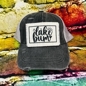 May include: A distressed charcoal gray baseball cap with a white mesh back. The front features a white rectangular patch with the words "lake bum" in black script and a sun graphic. The cap has a pre-curved bill.