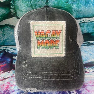 May include: A black and white trucker hat with a distressed look. The hat has a patch on the front that says "VACAY MODE" in rainbow colors.