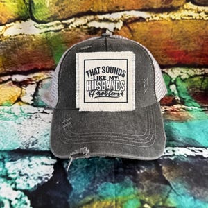 May include: A distressed gray trucker hat with a white mesh back. The front features a square patch with the text "THAT SOUNDS LIKE MY HUSBANDS PROBLEM" in black lettering. The hat has a curved brim and a worn appearance.