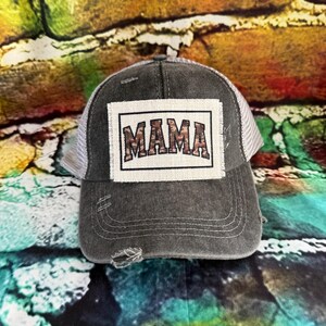May include: A distressed gray trucker hat with a white mesh back. The front features a rectangular patch with the word "MAMA" in a camouflage pattern. The hat has a pre-curved bill and a distressed look.