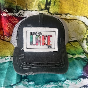 May include: Black and gray trucker hat with a distressed look. The hat has a patch on the front that says "Living on Lake Time."