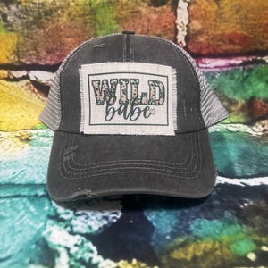 May include: A black and gray distressed trucker hat with a patch that says "WILD babe" in a leopard print font with a pink heart.