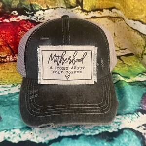 May include: A black and gray distressed trucker hat with a white patch that reads "Motherhood A Story About Cold Coffee" with a small heart symbol.