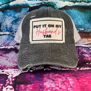 May include: Distressed charcoal gray trucker hat with a white mesh back. The front patch reads "PUT IT ON MY Husband's TAB" in white and pink lettering. The hat has a pre-curved bill and a distressed look.