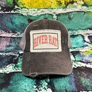May include: A distressed gray trucker hat with a white mesh back and a curved brim. The hat features a rectangular patch with the words "RIVER RAT" in red letters.