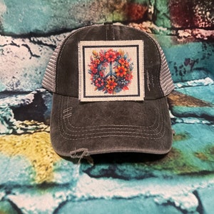 May include: A black and gray distressed trucker hat with a floral peace sign patch. The patch is a square with a white background and a black border. The peace sign is surrounded by colorful flowers.