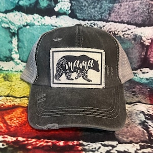 May include: A black and gray distressed trucker hat with a white patch featuring a black bear silhouette and the word "mama" in white lettering.