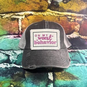 May include: A black and gray distressed trucker hat with a white patch that says "On my worst behavior" in pink lettering.