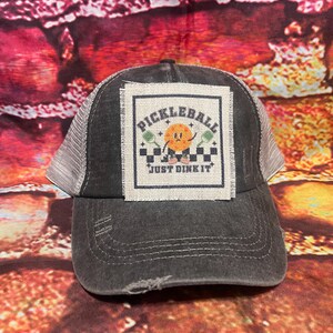 May include: A black and gray trucker hat with a white patch that says "Pickleball Just Dink It" and features a cartoon pickleball with a face.