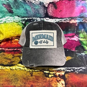 May include: A gray and white trucker hat with a distressed brim. The hat features a rectangular patch with the words "MERMAID off duty" in teal lettering and a seashell graphic.