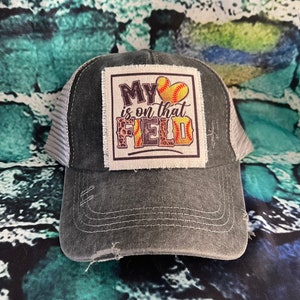 May include: A distressed gray baseball cap with a white mesh back. The front features a square patch with the text "My is on that FIELD" and a softball graphic. The hat has a pre-curved bill.