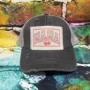 May include: A black and gray distressed trucker hat with a patch that says "MAMA" in red and white letters with a red cherry design.