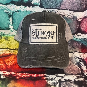 May include: A black and gray distressed trucker hat with a white patch that says "stronger than the storm" with a small heart.