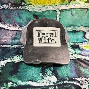 May include: A gray trucker hat with a mesh back and a distressed look. The front of the hat features a white rectangular patch with the words "Feral Wife" in black, block letters. The hat has a curved brim.