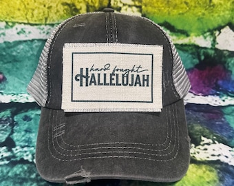 May include: A black and gray trucker hat with a distressed look. The hat has a patch on the front that says "hard fought HALLELUJAH".