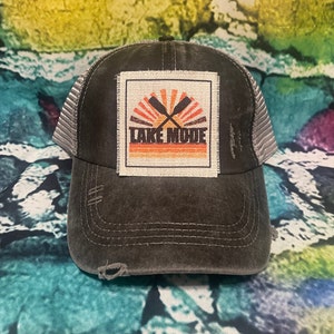 May include: A black and gray distressed trucker hat with a patch that says "Lake Mode" in white text with a sunset and two crossed oars in orange and red.