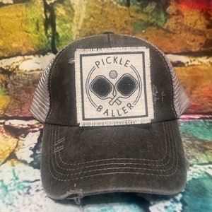 May include: A black and gray distressed trucker hat with a white patch that says "Pickle Baller" and features two pickleball paddles and a ball.