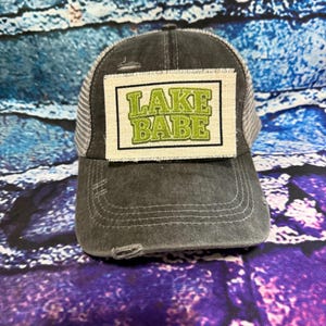 May include: A distressed gray trucker hat with a white mesh back and a curved brim. The hat features a rectangular patch with the words "LAKE BABE" in green, outlined in black, and set against a cream background. The hat is designed for casual wear.