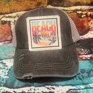 May include: A black and gray distressed trucker hat with a patch that says "Beach Bum" in rainbow colors with palm trees and a sunset.
