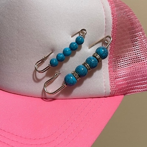 Może przedstawiać: Dwie turkusowe i srebrne broszki z zapinką typu safety pin z kryształkami. Broszki znajdują się na r&oacute;żowo-białej czapce z daszkiem.