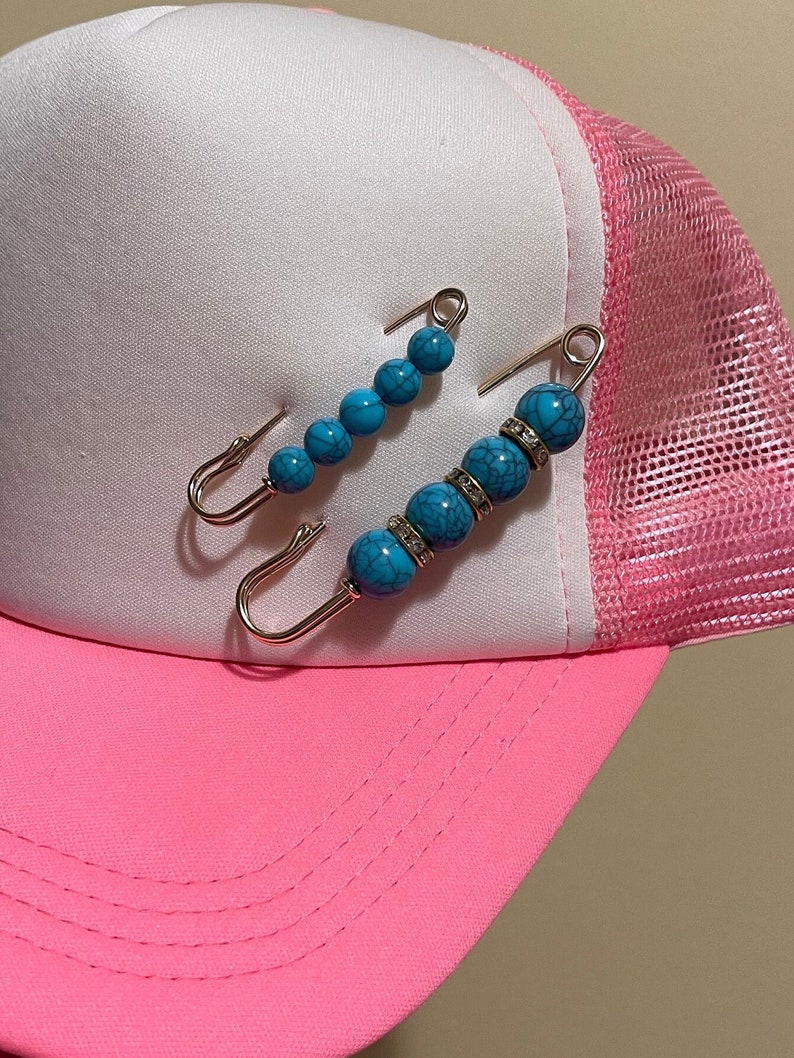 Może przedstawiać: Dwie turkusowe i srebrne broszki z zapinką typu safety pin z kryształkami. Broszki znajdują się na r&oacute;żowo-białej czapce z daszkiem.