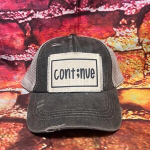 May include: A distressed black trucker hat with a beige mesh back and a curved brim. The hat features a rectangular patch with the word "continue" in black lettering.
