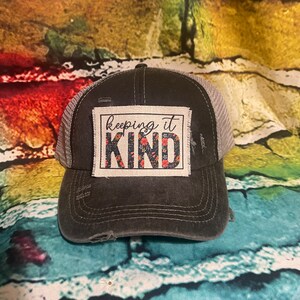May include: A black and gray distressed baseball cap with a white patch that says "keeping it KIND" in black lettering with a floral design in red, black, and white.