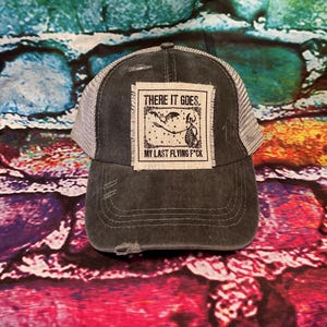 May include: A black and gray distressed trucker hat with a white patch that says "THERE IT GOES. MY LAST FLYING F*CK" with a skeleton graphic.