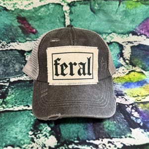 May include: A distressed gray trucker hat with a mesh back and a curved brim. The hat features a rectangular patch with the word "feral" in a gothic font.