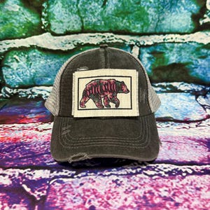 May include: A charcoal gray distressed baseball cap with a mesh back and a rectangular patch on the front. The patch features a black outline of a bear filled with a floral pattern and the word mama written in a cursive pink script font.
